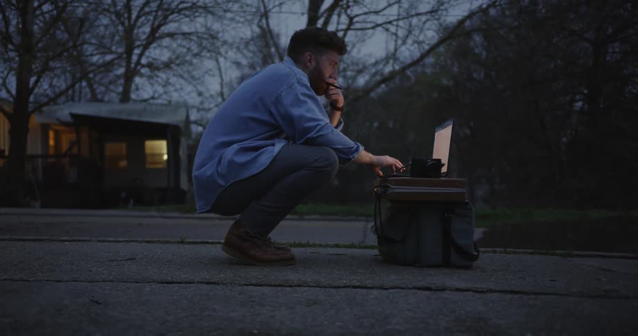Man squatting outside working on a laptop propped up on a suitcase and bag