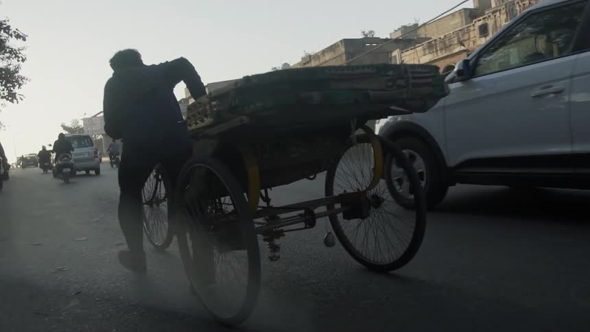 A man walking and pushing a tricycle on a street with vehicles with buildings the sky and clouds in the background