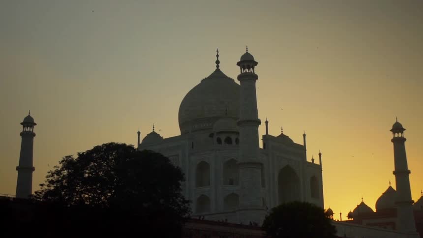 A white temple or mosque with a dome against a sunset with a birding flying in the sky and trees on one side