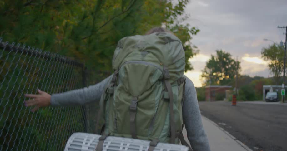A young woman walking down a neighborhood street with a backpack on runs her hand along a fence as she walks