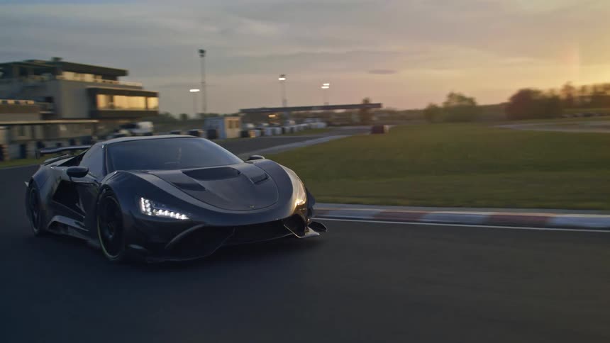 A sports car driving on a track with buildings trees the sky and clouds in the background