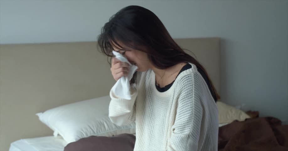 A woman sitting on a bed crying wiping her face with a tissue