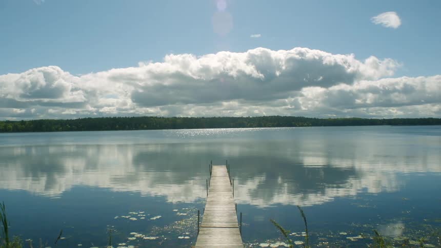 A wooden pier reaches out into a lake reflecting the sunny daytime sky