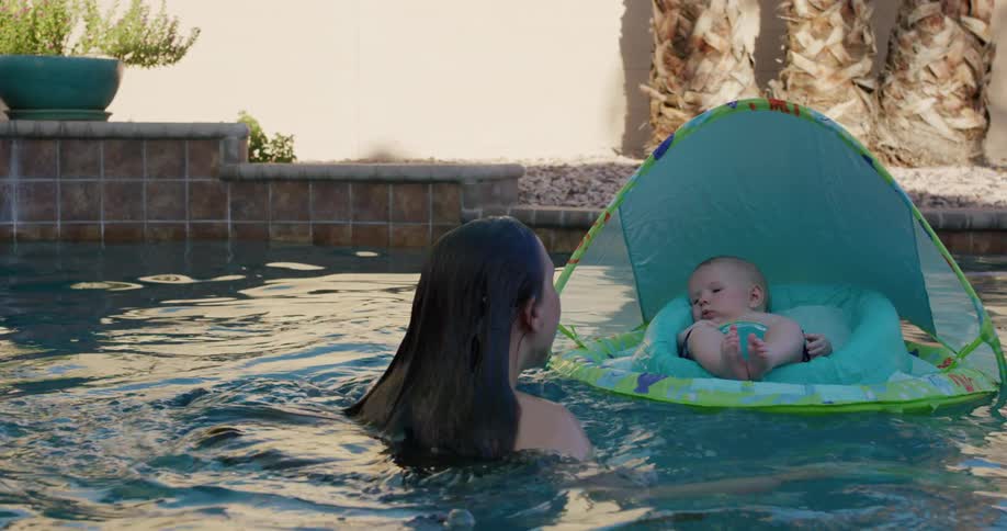 Baby floats on float in pool laughing when its mother emerges from pool water nearby