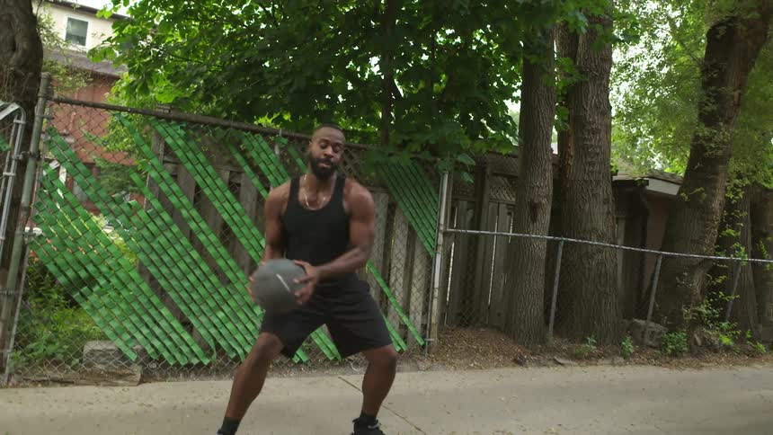 A young woman and a man pass an exercise ball back and forth between them as they exercise in an alleyway in a neighborhood during the day