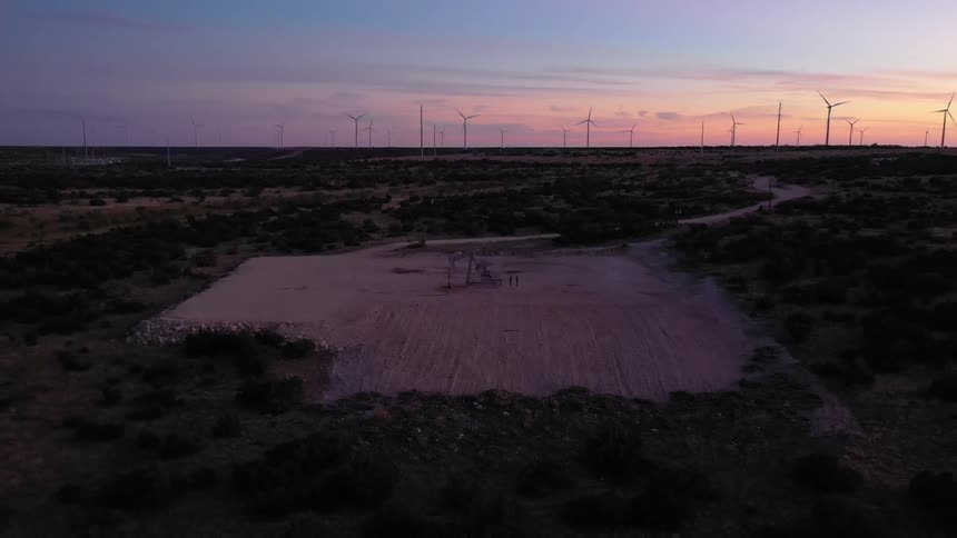 An oil well surrounded by sand with wind turbines lined up in the distance with a setting sun