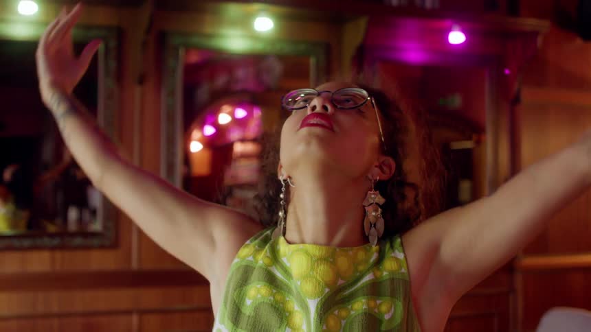 A young woman wearing glasses and elaborate ear rings dances indoors during the day