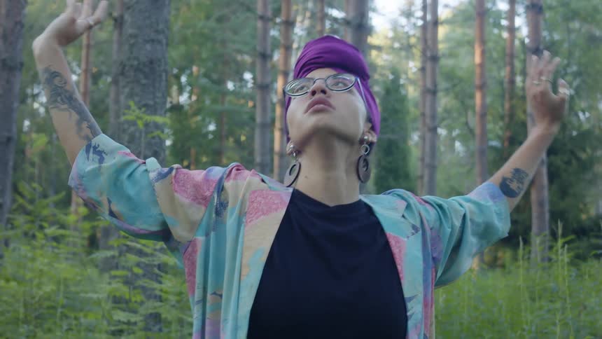 A woman with glasses dancing in the forest with trees the sky and clouds in the background