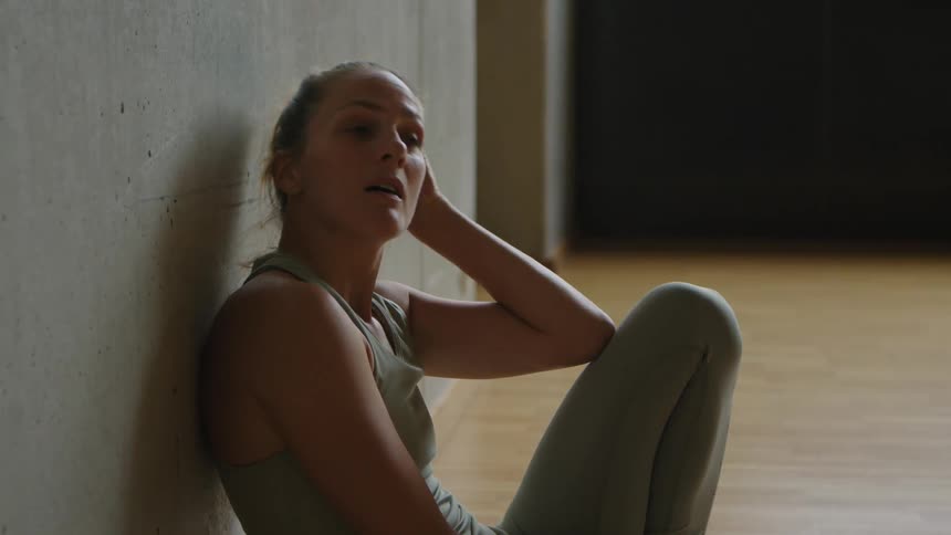 Woman with a sport top and leggings sits and leans against the wall after workout in the gym