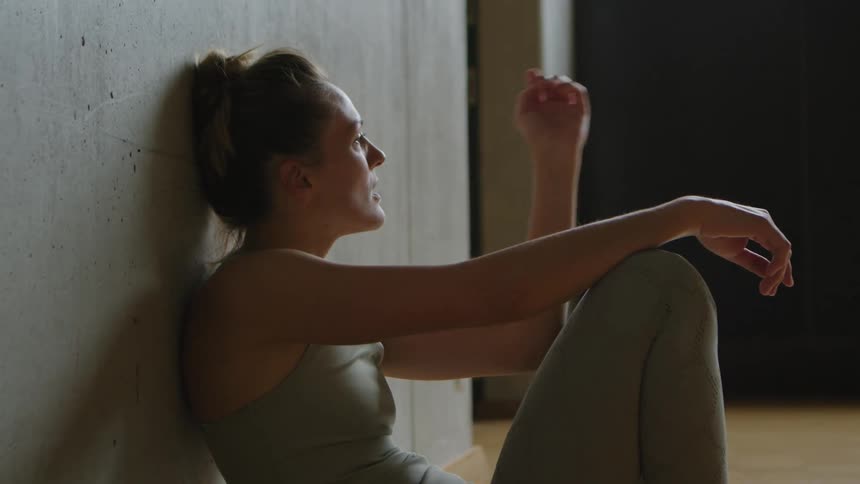 Woman with a sport top and leggings sits and leans against the wall after workout in the gym