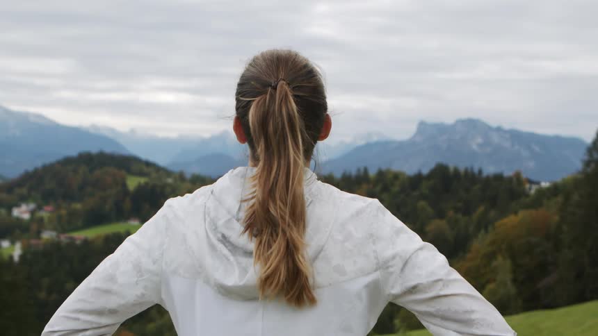 Woman runs over to the edge of a hillside and looks off at a mountain range