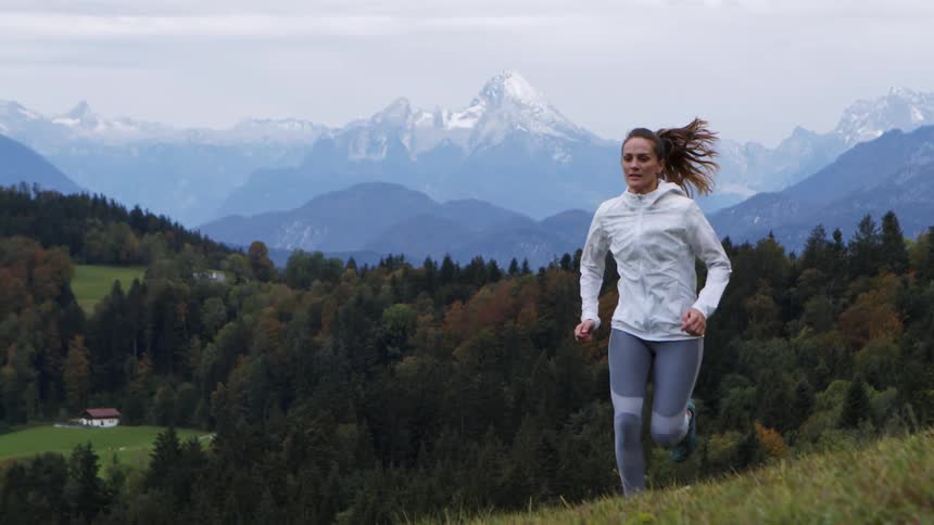 Woman jogs up grass covered slope near forest backed with mist covered mountains