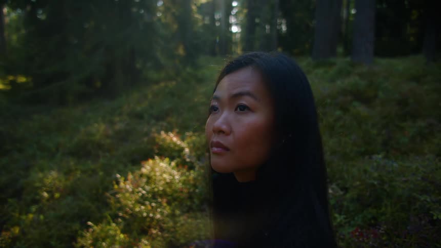 An asian woman stands outside in a forest looking around at sunset