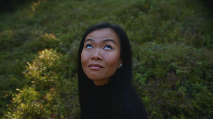 Woman with dark straight hair and a black sweater looks up at the sky in the forest