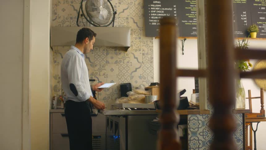 A man in apron uses a tablet while standing in a kitchen before returning it to its stand above the oven