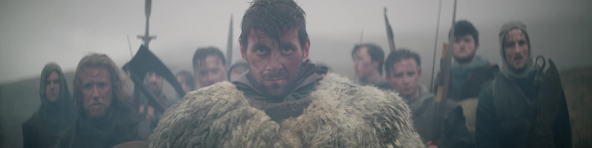 A man wearing a fur cloak stares straight ahead while standing in front of a group of men holding weapons