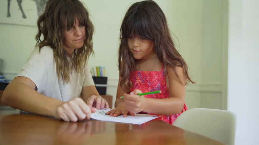 A woman sits next to her daughter and hands her a pencil to write on a paper