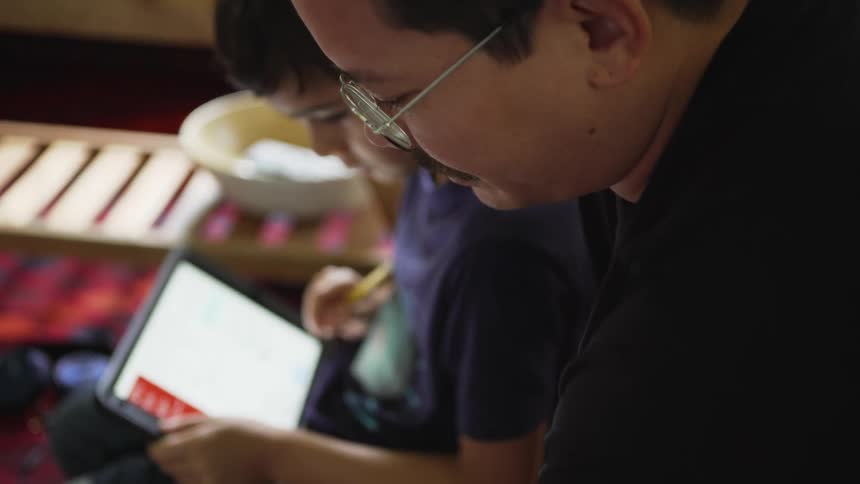A little boy sits with his fathers and they look at a tablet and then the boy erases text on a paper