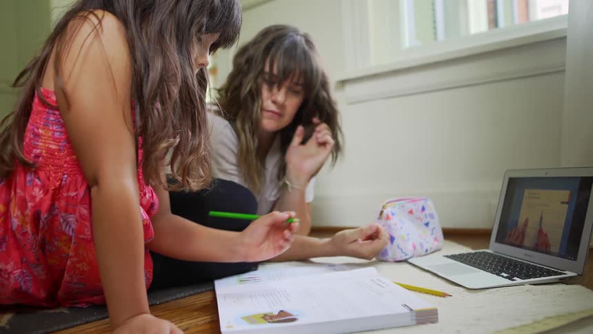 A mother helps her daughter complete homework on paper while sitting in front of a laptop in the floor indoors