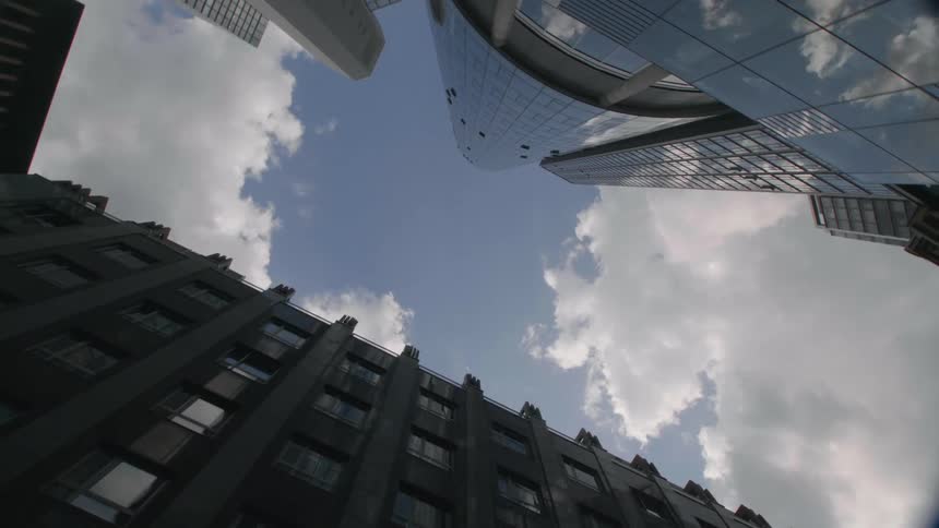 Fluffy clouds in blue sky are framed and reflected by tall buildings and a few trees