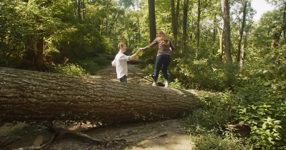 Man with a white shirt and jeans holding the hand of a woman wearing a sweater and jeans helps her walk over a tree trunk in the woods