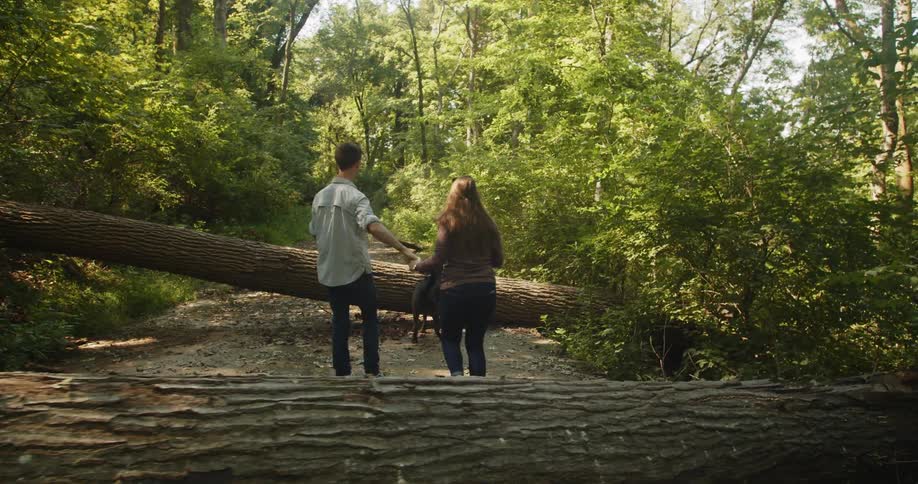 Man holding the hand of a woman helps her walk over a tree trunk in the forest