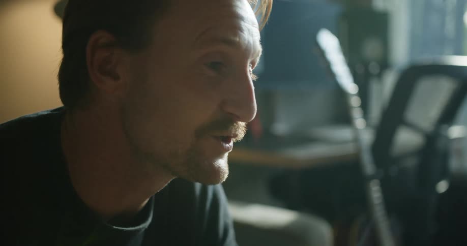 A man is talking a throwing a kiss as he sits indoors in front of a piano and guitar