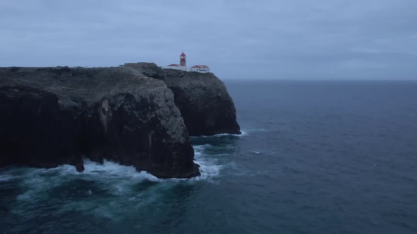 Lighthouse building on the edge of a cliff with waves splashing up against the cliff side