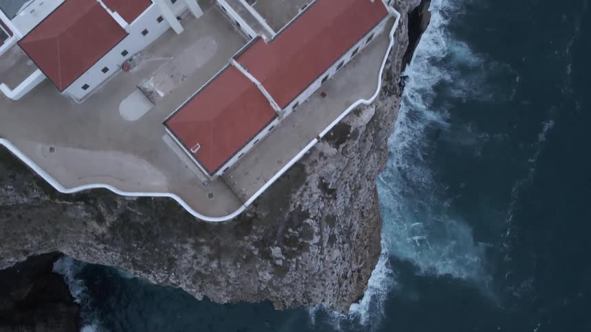 Waves crashing up against the side of a ocean side cliff with a large building structure and a lighthouse on the coast