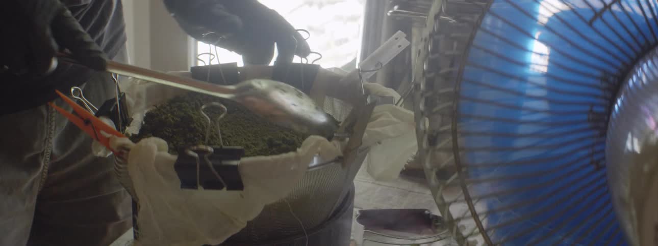 A person is using a spoon to press down items in a container with a fan blowing on them inside