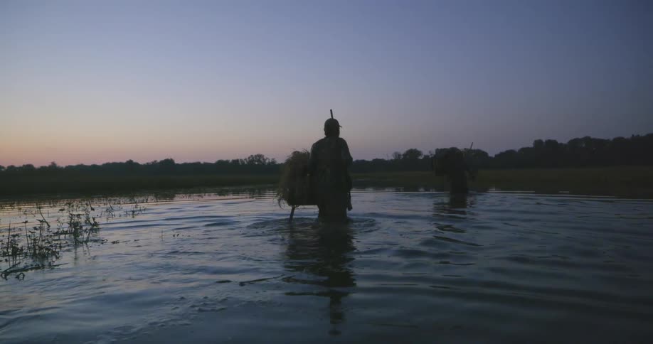 Men carrying rifles wade through water toward tree covered shore as sky at horizon remains tinted pink