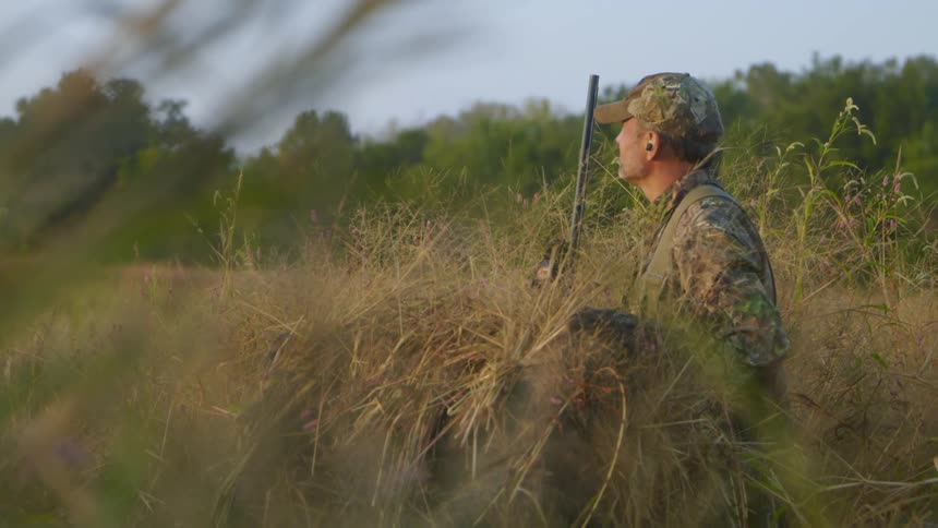Hunter stands in a grassy field with his gun and scans the area with his eyes