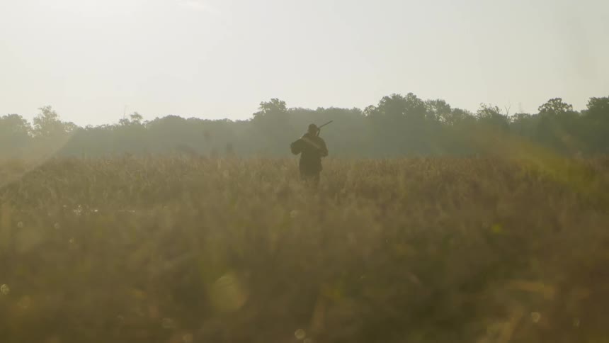 Man carrying a long gun walking through a puddle in an open grass field