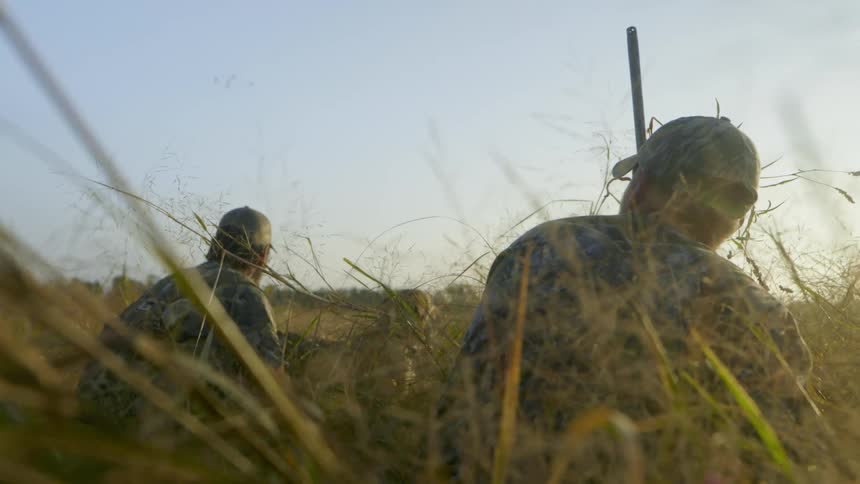 Two men wear camouflage while sitting in a field and one uses a bird call