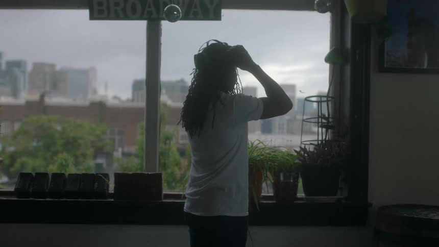 Man in dark room walks across and puts on vr headset with plants and a window behind him where you can see a city skyline