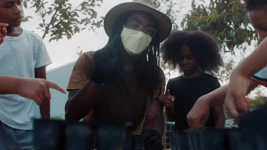 A man wearing a sun hat helps children plant seeds in small containers outdoors