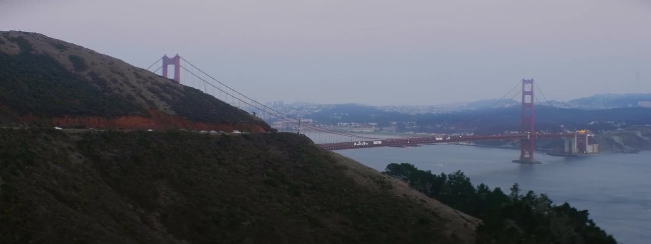 Cars drive along suspension bridge over the ocean with hills before and after in the sunrise