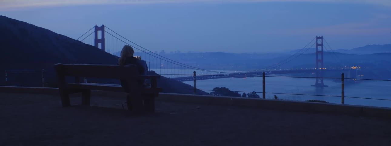 A woman sits on a bench and takes a photo with her cell phone while looking at the water and skyline