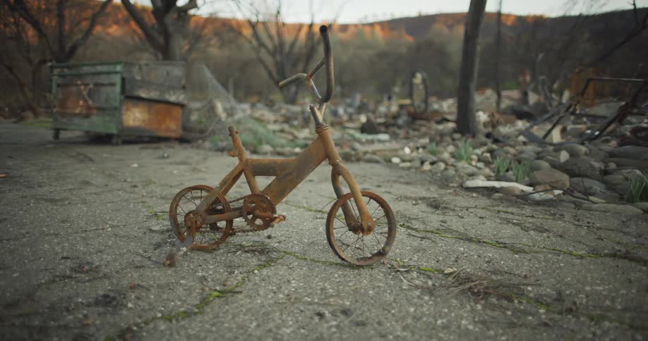 Old rusty tricycle on broken road with moss missing seat in front of rocks trees and trash bin with trees in the distance