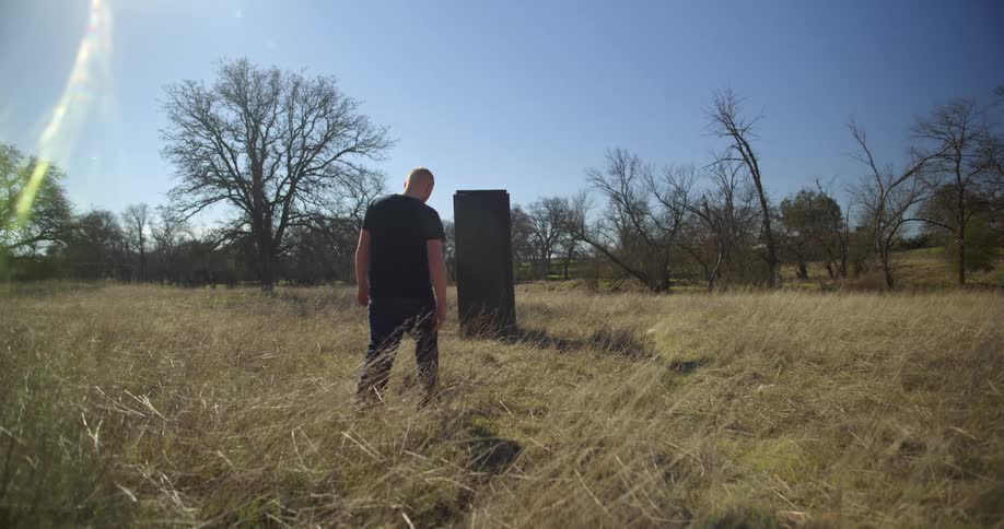 A man wearing all black walks outdoors through a field with long grass towards a tall black object