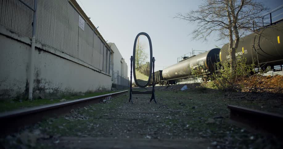 Oval standing mirror on a broken train track next to a train with a carrier tank