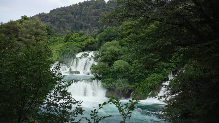 A waterfall pours water into a river that runs through a forested area