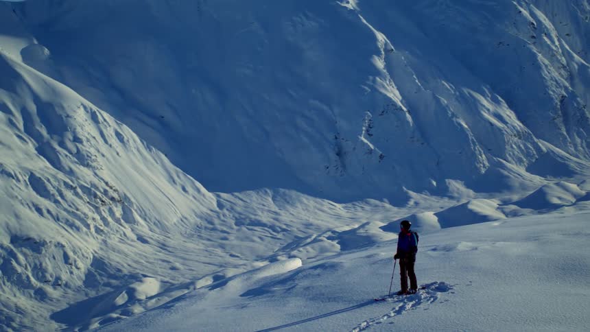Person standing at the top of a snowy mountain cliff at day time looking down at a slope descending into a valley
