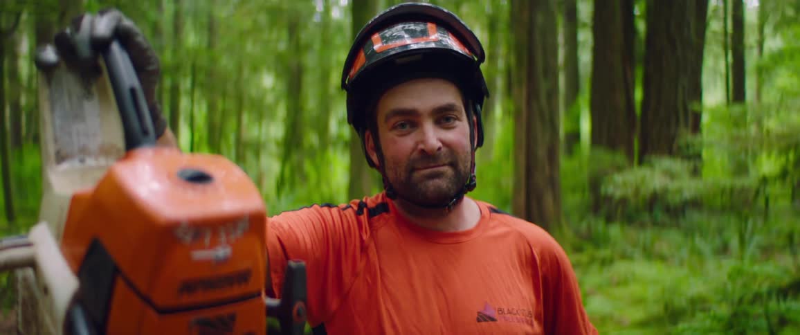 A lumberjack in personal protective equipment poses with his chainsaw while looking into the camera in a forest during the day