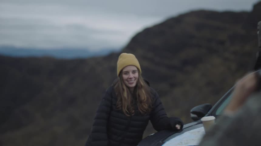 Man takes picture of a woman leaning on a car with hills in the background