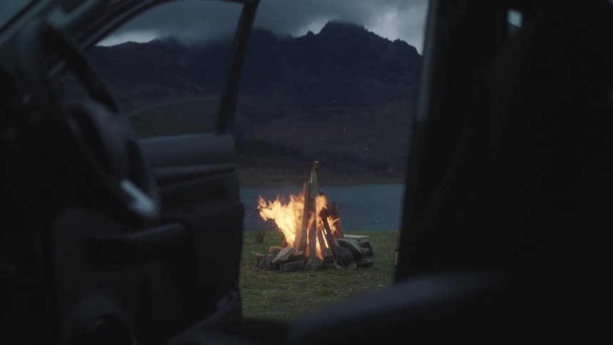 About 15 feet away from the passenger side of a car a bonfire is blazing in front of a lake with mountains in the background