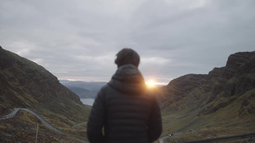 Man standing at the edge of a cliff overlook in a mountain range at sunset watching the sun set