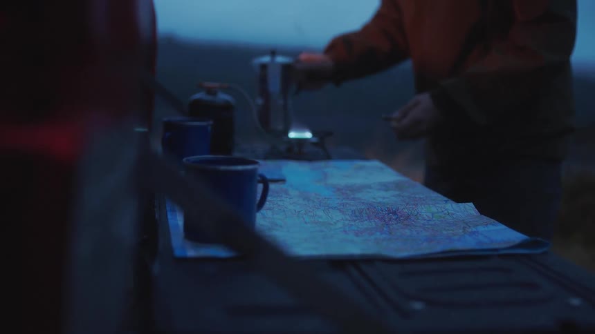 A man lights a burner for a coffee maker outdoors in a camping setting at sunrise