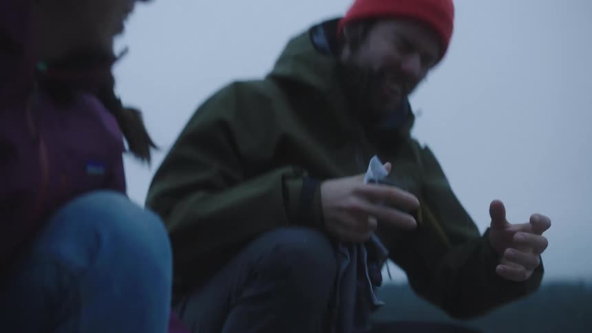 Male and female wearing jackets and hats washing camping dishes outside in a small area of a lake or stream