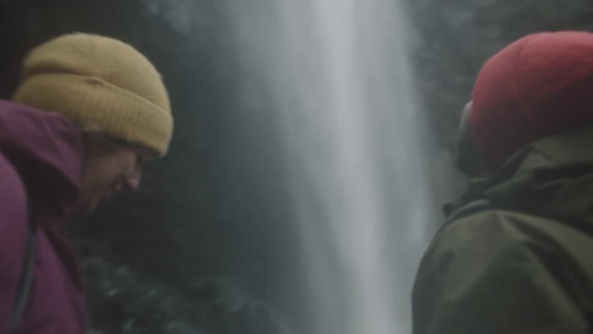 A man and woman kiss and congratulate each other as they approach the base of a waterfall in the wilderness during the day
